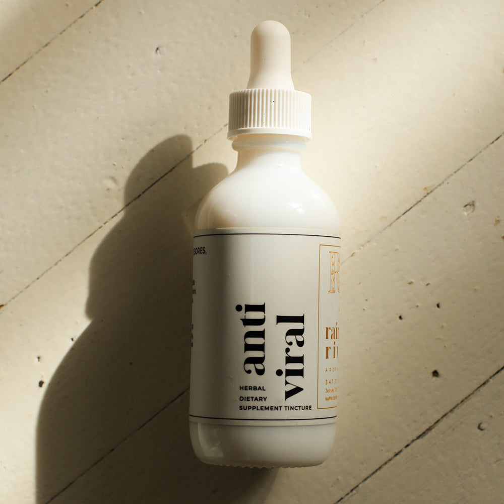White tincture droplet bottle with a minimal white label with black lower-case serif text saying "anti viral". The bottle sits on a light wooden background with a drop shadow going to the bottom left.