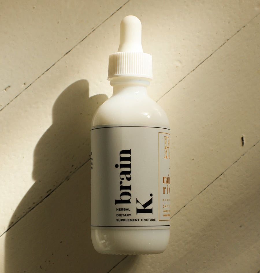 White tincture droplet bottle with a minimal white label with black lower-case serif text saying "brain K." The bottle sits on a light wooden background with a drop shadow going to the bottom left.