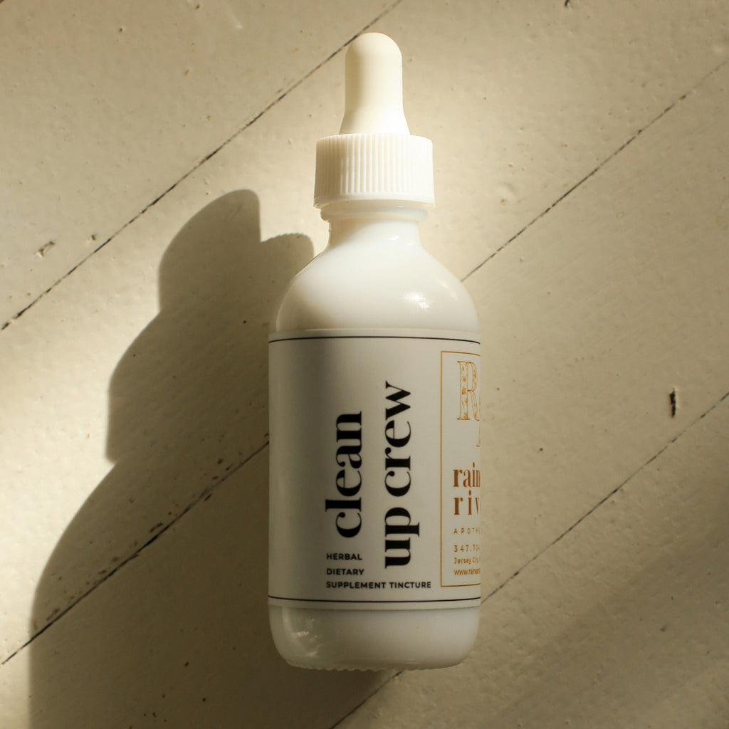 White tincture droplet bottle with a minimal white label with black lower-case serif text saying "clean up crew". The bottle sits on a light wooden background with a drop shadow going to the bottom left.