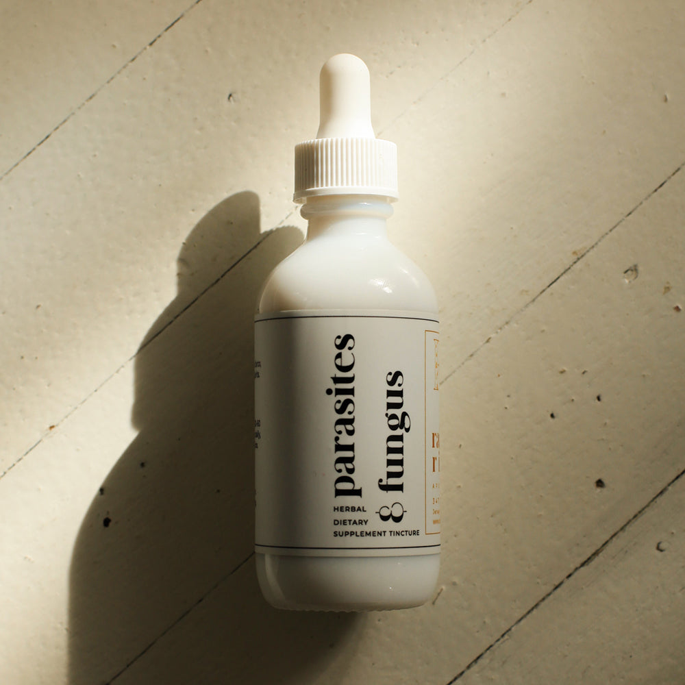 White tincture droplet bottle with a minimal white label with black lower-case serif text saying "parasites & fungus". The bottle sits on a light wooden background with a drop shadow going to the bottom left.